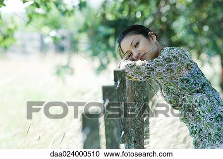 Stock Image - Young woman leaning against rural fence, head resting on arms, smiling at camera. Fotosearch