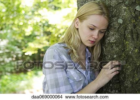 Picture - Young woman leaning against tree, looking sad. Fotosearch