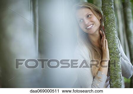 Stock Image - Young woman leaning against tree trunk, focus on background. Fotosearch