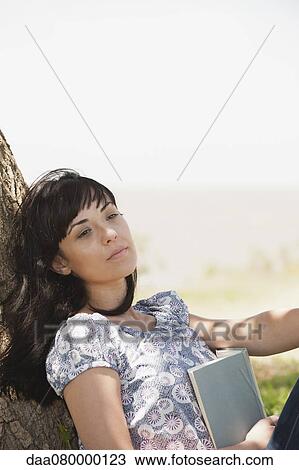 Stock Image - Young woman leaning against tree trunk holding book. Fotosearch