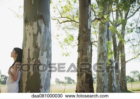Stock Image - Young woman leaning against tree trunk. Fotosearch
