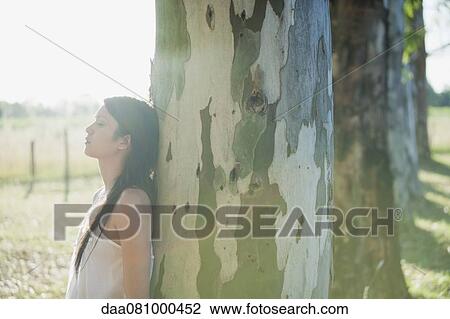 Stock Image - Young woman leaning against tree trunk. Fotosearch