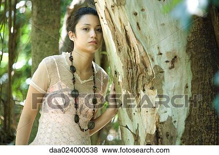 Stock Photo - Young woman leaning against tree trunk, looking at camera. Fotosearch