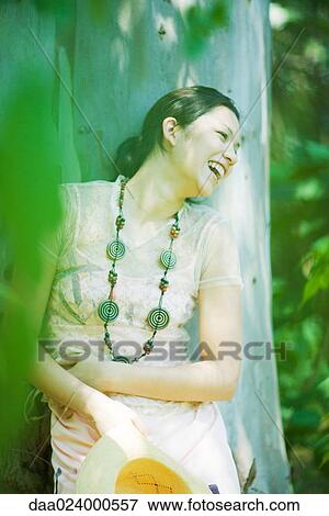 Young woman leaning against tree trunk, laughing, looking away View Large Photo Image Stock Photo - Young woman leaning against tree trunk, laughing, looking away. Fotosearch