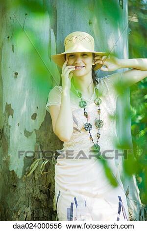 Young woman leaning against tree trunk, smiling at camera, view through foliage View Large Photo Image Stock Photograph - Young woman leaning against tree trunk, smiling at camera, view through foliage. Fotosearch
