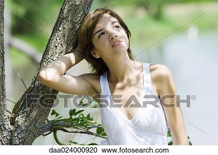 Stock Image - Young woman leaning against tree with hand behind head, looking up. Fotosearch