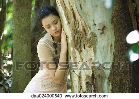 Stock Image - Young woman leaning head against tree trunk, eyes closed. Fotosearch