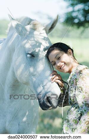 Stock Photo - Young woman leaning her head against horse, smiling. Fotosearch
