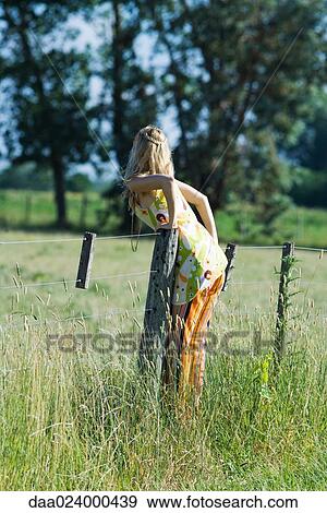 Stock Photo - Young woman leaning over rural fence, rear view. Fotosearch