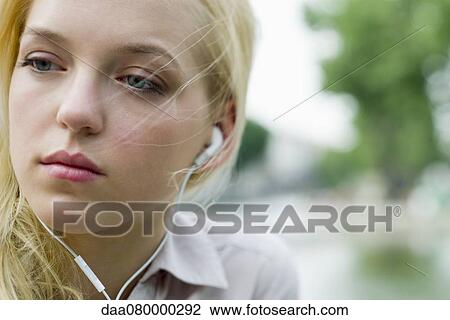 Stock Image - Young woman listening to earphones, looking away, portrait. Fotosearch