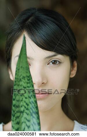 Stock Image - Young woman looking at camera with leaf covering one eye, portrait. Fotosearch
