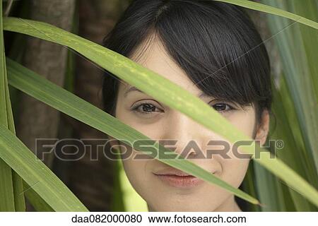 Stock Image - Young woman looking through foliage, portrait. Fotosearch
