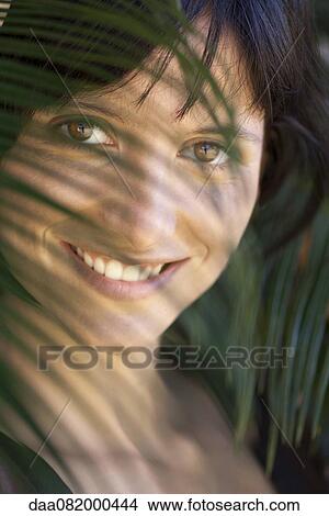 Young woman looking through palm leaf, smiling, portrait View Large Photo Image Picture - Young woman looking through palm leaf, smiling, portrait. Fotosearch