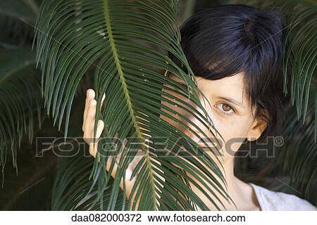Stock Image - Young woman looking through palm leaf, portrait. Fotosearch