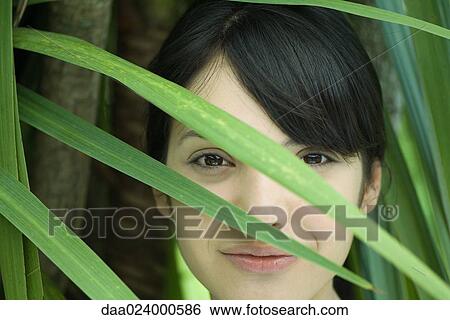 Stock Photograph - Young woman looking through palm leaf, smiling at camera, portrait. Fotosearch