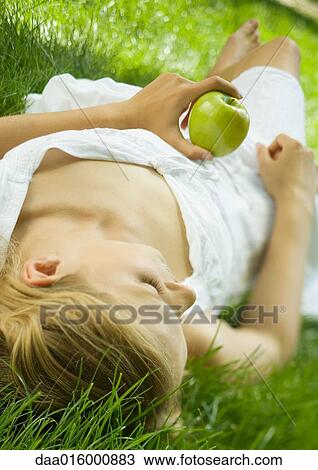 Young woman lying in grass, holding apple View Large Photo Image Stock Image - Young woman lying in grass, holding apple. Fotosearch