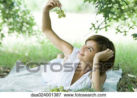 Stock Photo - Young woman lying on grass, holding up grapes. Fotosearch