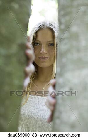 Stock Photo - Young woman peering out from behind tree trunks. Fotosearch