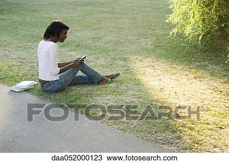 Stock Image - Young woman relaxing in park, text messaging with cell phone. Fotosearch