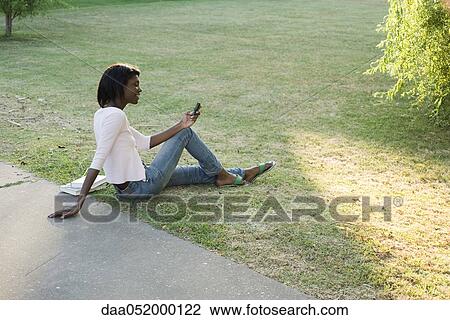 Stock Image - Young woman relaxing in park using cell phone. Fotosearch