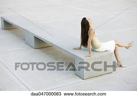 Stock Image - Young woman relaxing on bench in city square, high angle view. Fotosearch