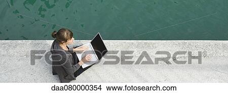 Picture - Young woman sitting beside canal, using laptop computer. Fotosearch