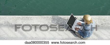 Stock Photography - Young woman sitting beside canal, using laptop computer. Fotosearch