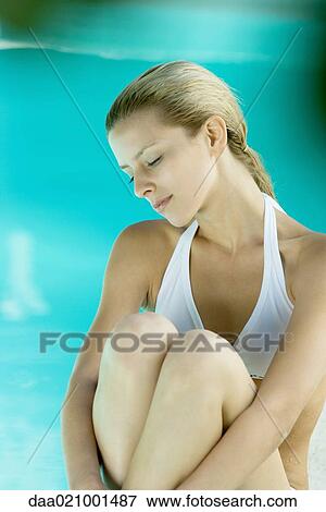 Stock Photo - Young woman sitting by edge of pool with knees up. Fotosearch