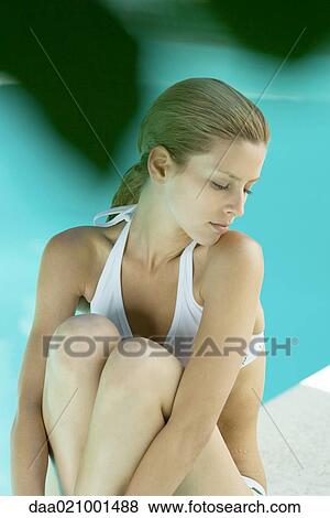 Stock Photo - Young woman sitting by edge of pool with knees up. Fotosearch