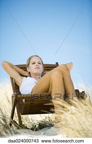 Stock Image - Young woman sitting in deck chair, in dunes. Fotosearch