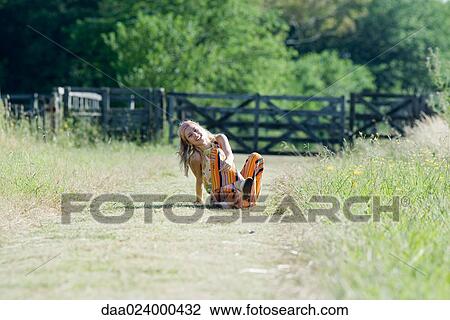 Stock Image - Young woman sitting in rural field with knees up, smiling at camera. Fotosearch