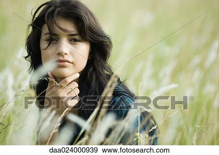 Young woman sitting in tall grass with hand under chin, looking at camera View Large Photo Image Stock Photo - Young woman sitting in tall grass with hand under chin, looking at camera. Fotosearch