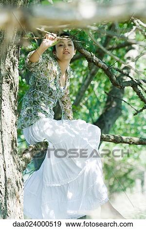 Stock Photo - Young woman sitting in tree, looking at camera. Fotosearch