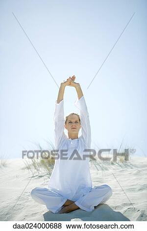 Stock Photo - Young woman sitting on dune, stretching arms over head. Fotosearch