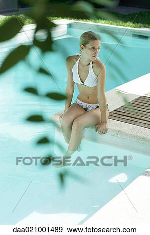 Stock Photo - Young woman sitting on edge of pool, full length, blurred leaves in foreground. Fotosearch