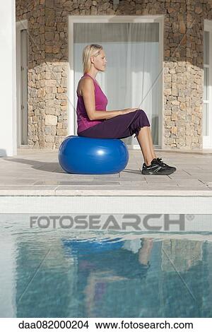 Picture - Young woman sitting on fitness ball at poolside. Fotosearch