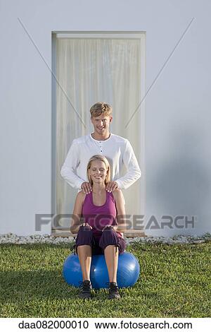 Stock Image - Young woman sitting on fitness ball, boyfriend giving her massage. Fotosearch