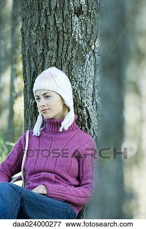 Stock Photo - Young woman sitting on ground, leaning against tree, wearing knit hat and sweater. Fotosearch