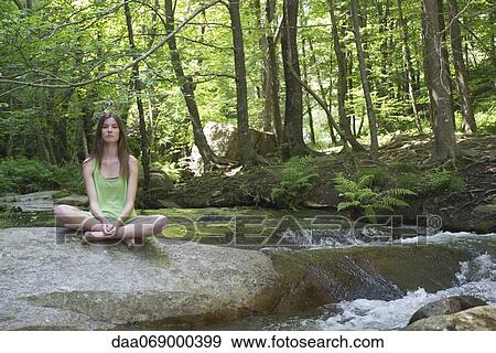 Stock Photo - Young woman sitting on rock beside stream. Fotosearch