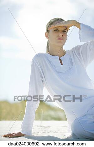 Young woman sitting on sand, shading eyes and looking out of frame View Large Photo Image Stock Image - Young woman sitting on sand, shading eyes and looking out of frame. Fotosearch