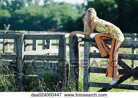 Young woman sitting on top of wooden fence, leaning on elbow, looking away View Large Photo Image Stock Photo - Young woman sitting on top of wooden fence, leaning on elbow, looking away. Fotosearch