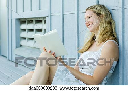 Young woman sitting outdoors, reading book, smiling View Large Photo Image Stock Image - Young woman sitting outdoors, reading book, smiling. Fotosearch