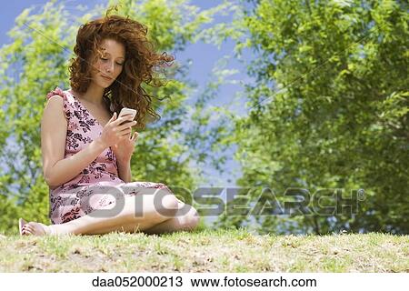 Stock Image - Young woman sitting outdoors, text messaging. Fotosearch