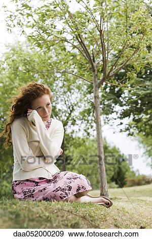 Stock Photo - Young woman sitting outdoors, wrapping herself in cardigan. Fotosearch
