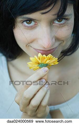 Stock Photo - Young woman smelling flower, smiling at camera, cropped, close-up. Fotosearch