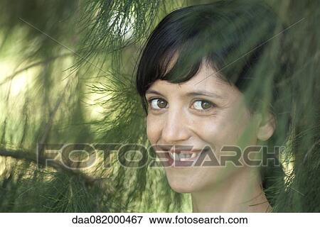 Young woman smiling through pine branches, portrait View Large Photo Image Stock Photo - Young woman smiling through pine branches, portrait. Fotosearch