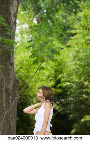 Stock Image - Young woman standing beside tree, smiling, side view. Fotosearch
