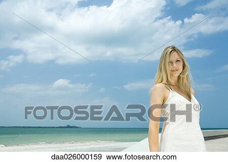 Stock Photo - Young woman standing by the beach, hair tousled by wind, smiling at camera. Fotosearch
