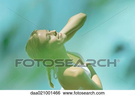 Stock Photograph - Young woman standing in bikini with hand behind head, eyes closed, high angle view, blur in foreground. Fotosearch