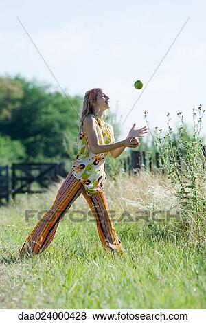 Stock Photo - Young woman standing in field with legs apart, juggling apples. Fotosearch